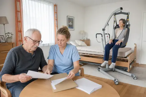 „Patientenlifter – Unterlagen“: Eine Angehörige und eine pflegebedürftige Person prüfen gemeinsam Pflegeunterlagen an einem Tisch.